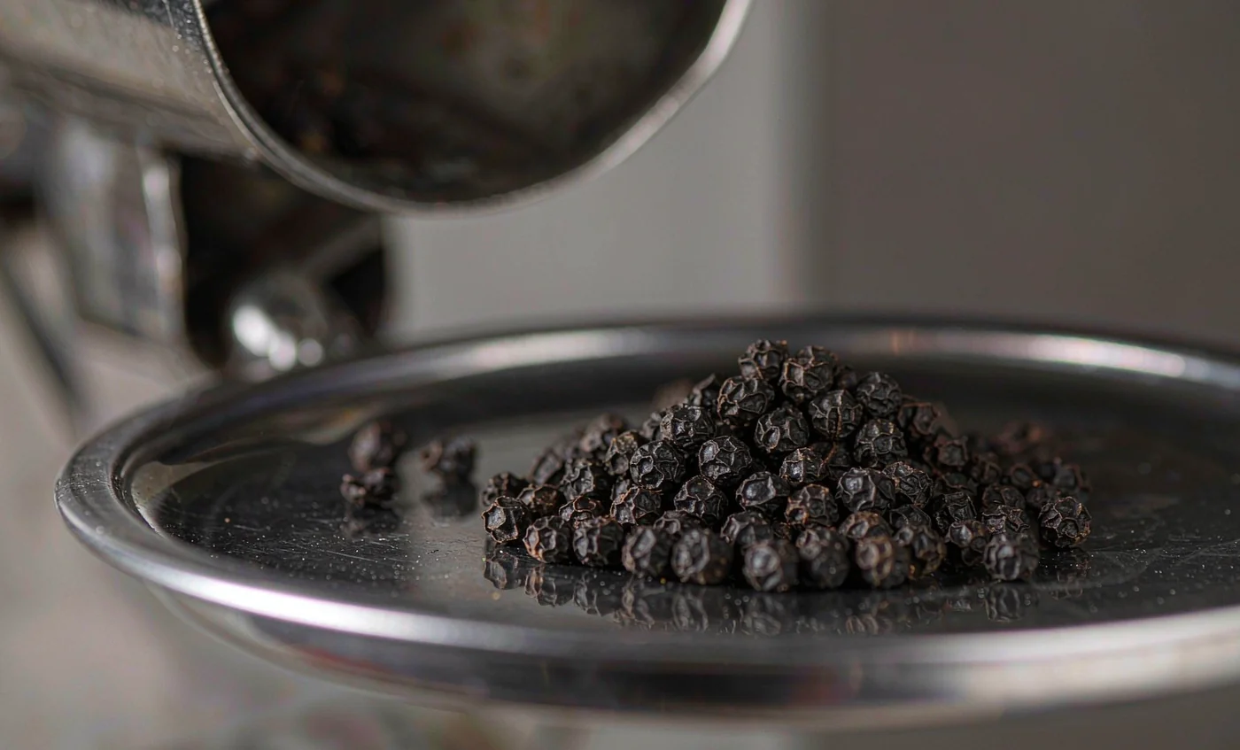 Raw black pepper corns on a stainless steel tray in front of an industrial grinder feed hopper